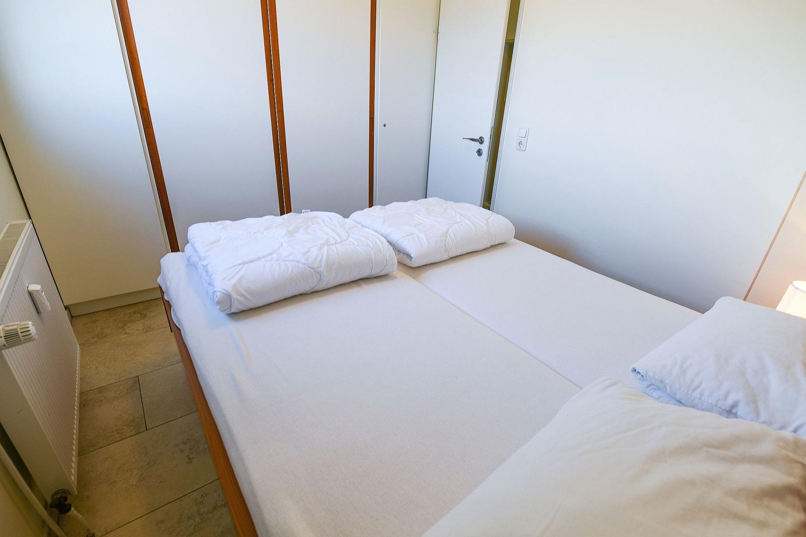 Zimmer mit Doppelbett, weißen Bettwäsche und Holzrahmen. Hintergrund: Schrank und Tür.