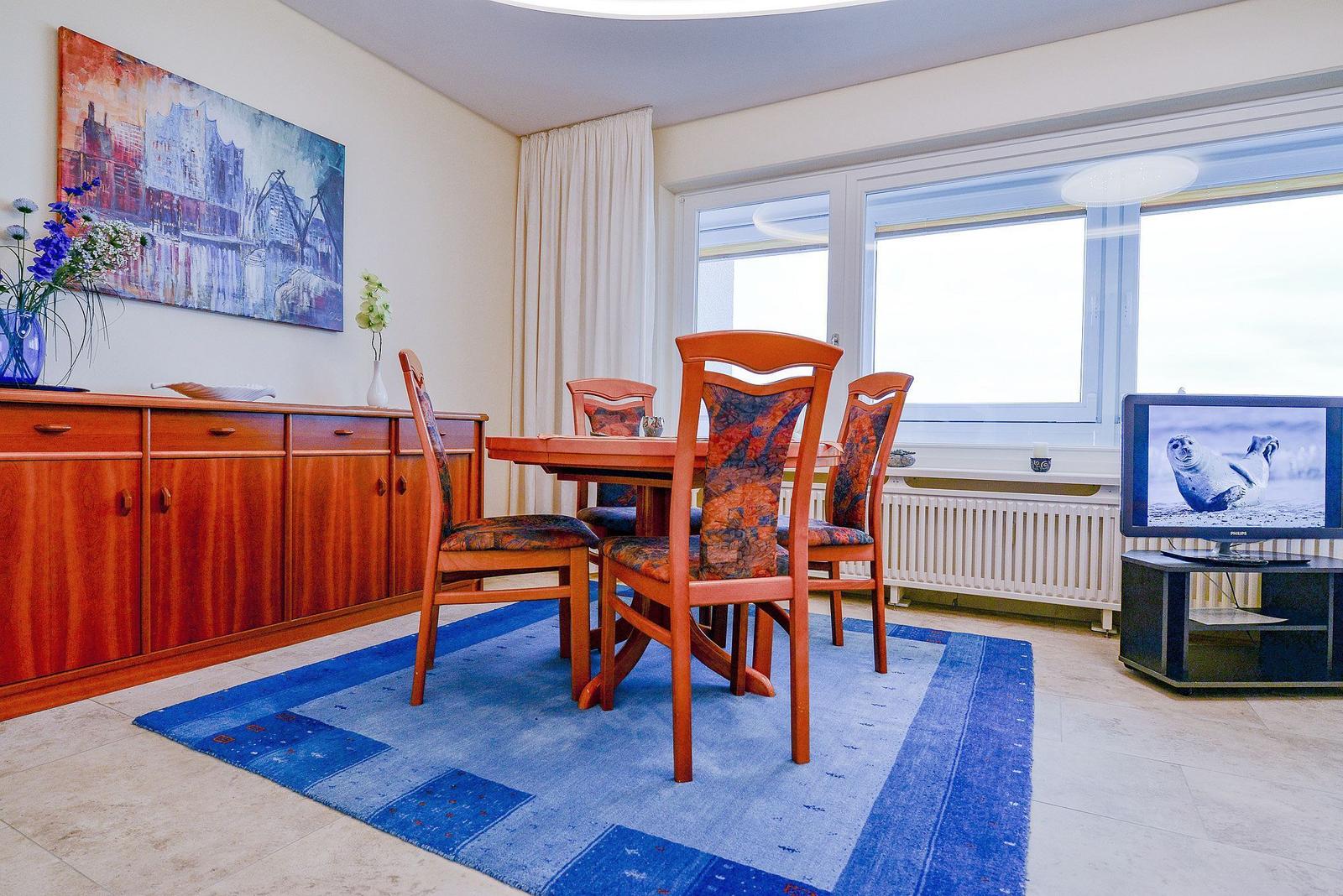 Esszimmer mit Holztisch, Stühlen und blauem Teppich. Fenster, TV und Wandbild sind sichtbar.