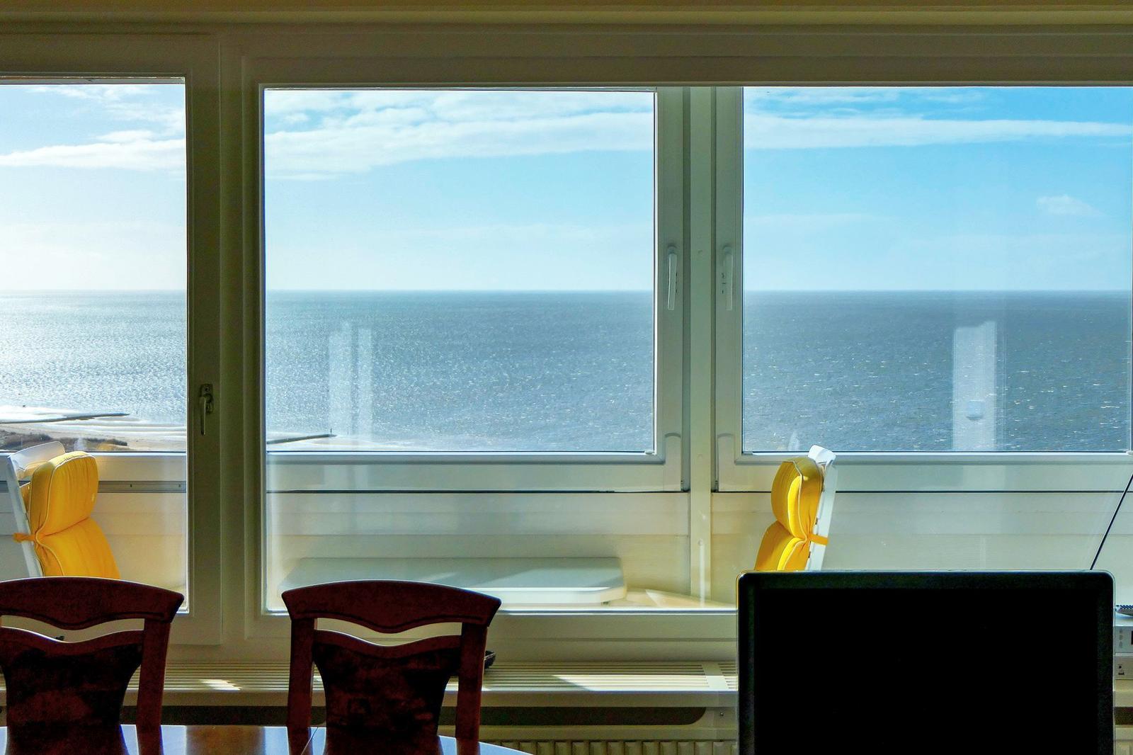 Großes Fenster mit Meerblick. Gelbe Sessel und Holzstühle vor dem Fenster.