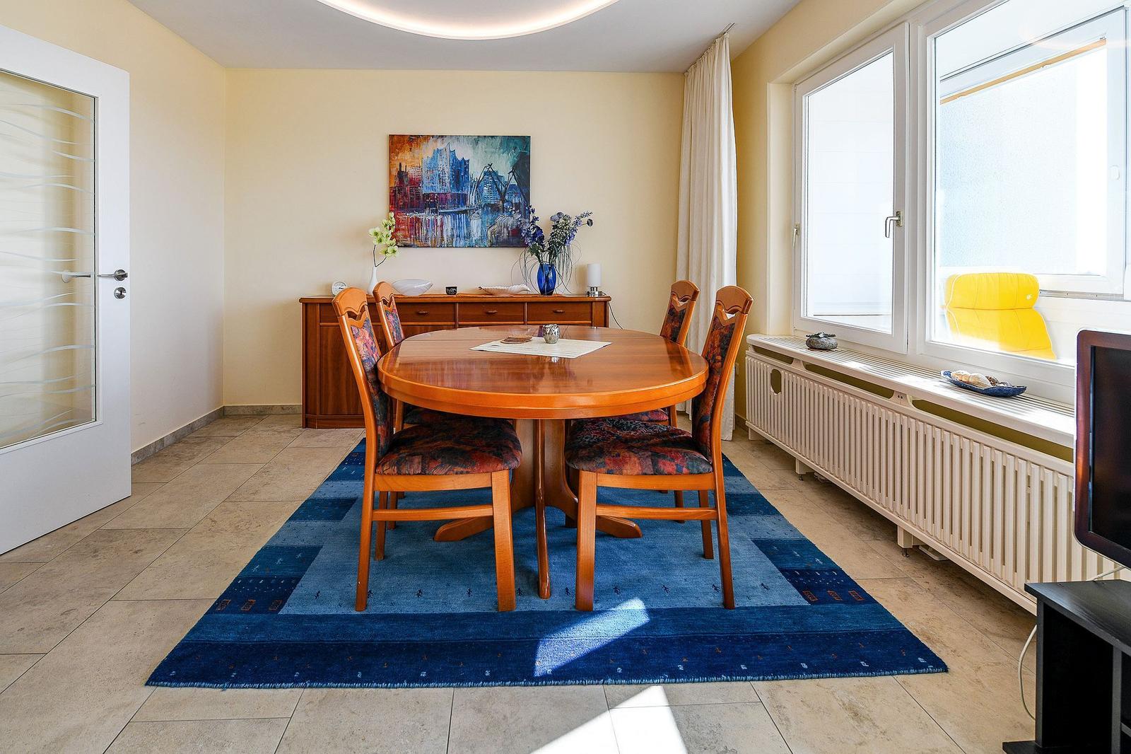 Esszimmer mit runden Tisch, vier Stühlen und blauem Teppich. Fenster und Wandbild sichtbar.