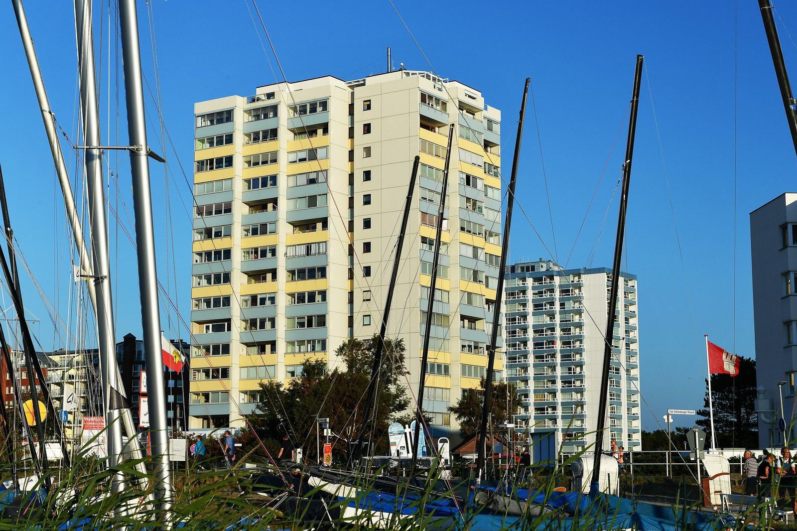 Hochhaus mit gelben und grauen Balkonen, umgeben von Segelyachten und blauem Himmel.