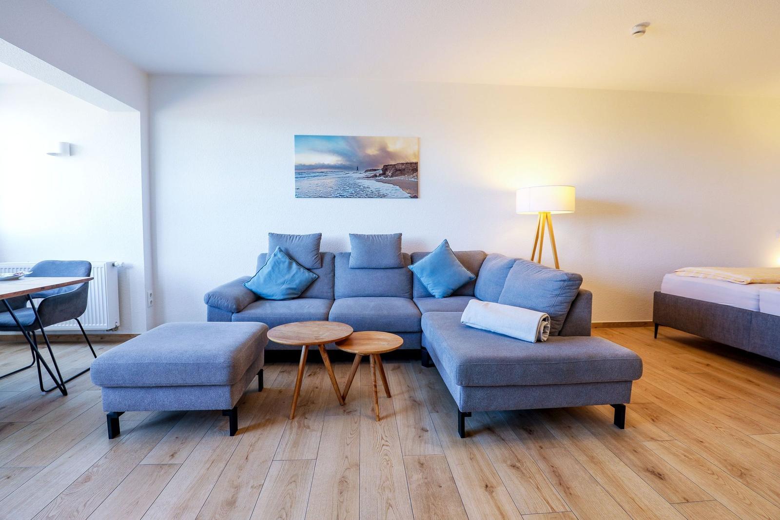 Wohnbereich mit Sofa, Tisch und Bett. Wandbild zeigt Strand.