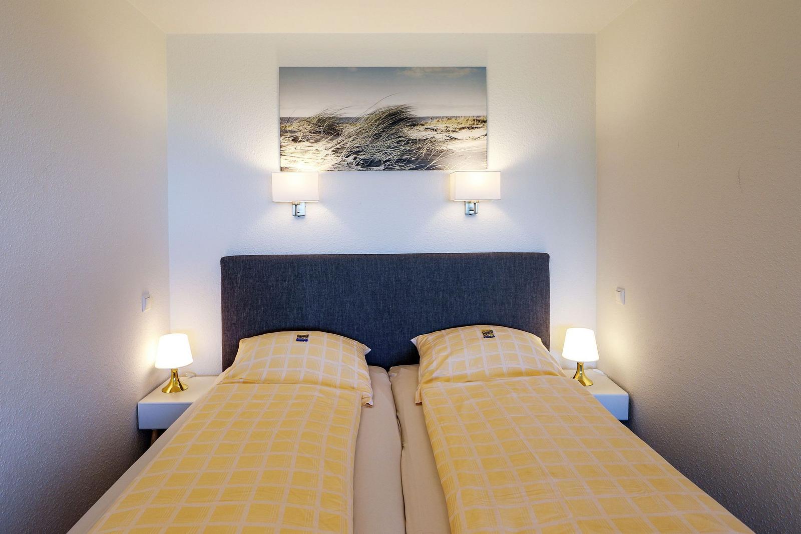 Zimmer mit Doppelbett, Nachttischen und Wandbild. Deckenleuchten und Kissen mit Muster.