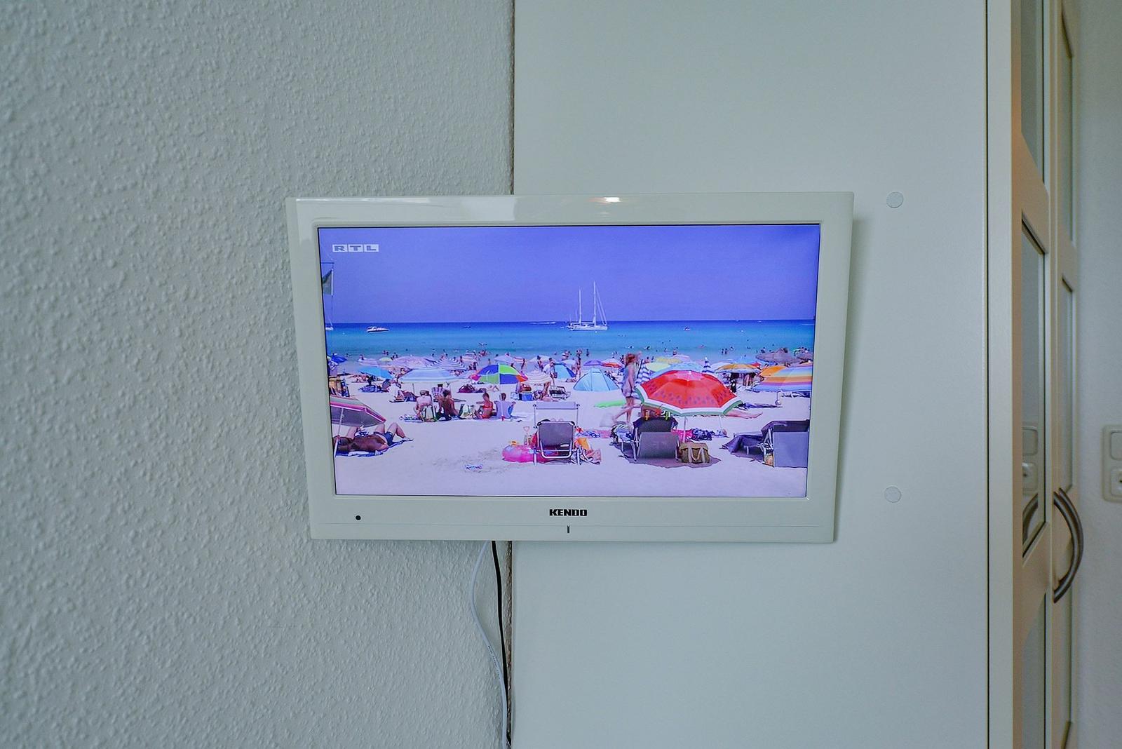 An der Wand montierter Fernseher zeigt einen Strand mit Sonnenschirmen und Booten.