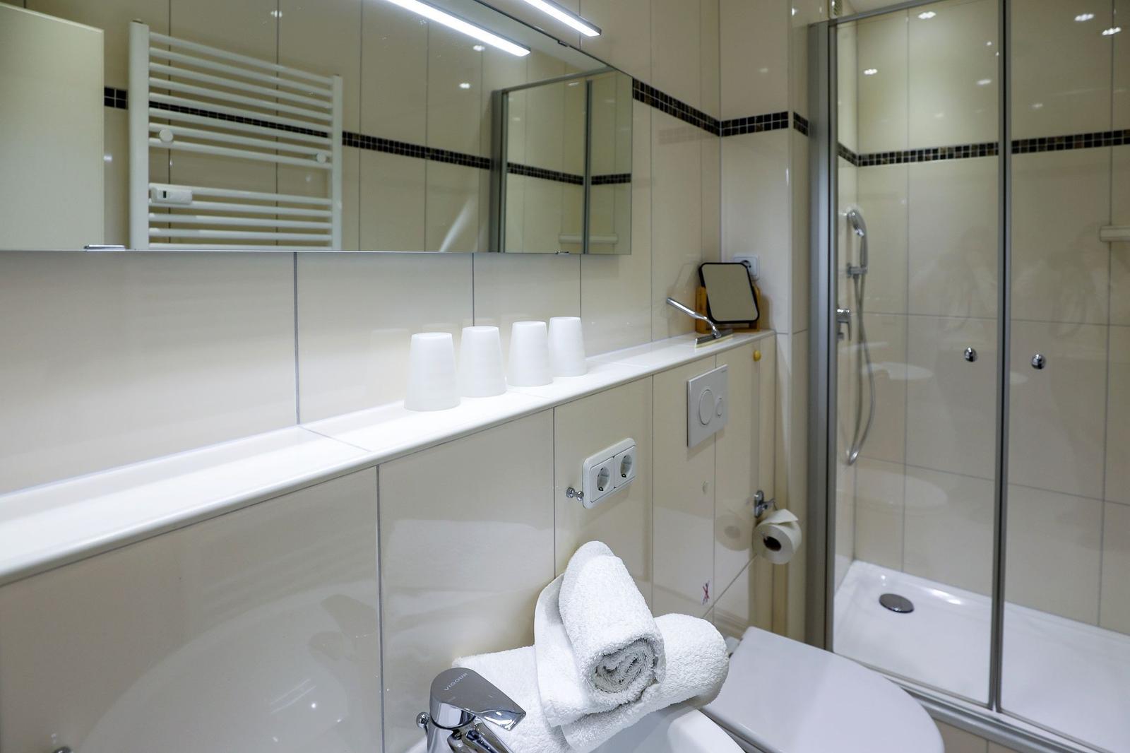 Badezimmer mit Dusche, Spiegel und Toilette. Weiße Fliesen und Handtücher.