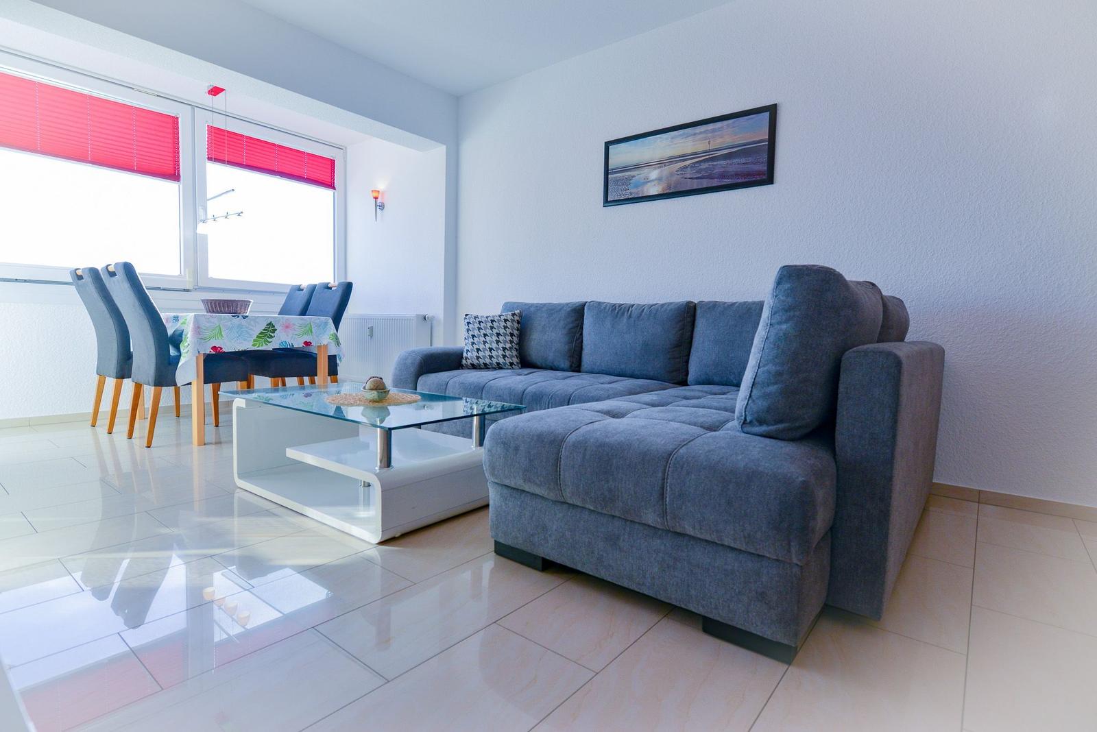 Wohn- und Essbereich mit grauem Sofa, Glas-Tisch und roten Jalousien.