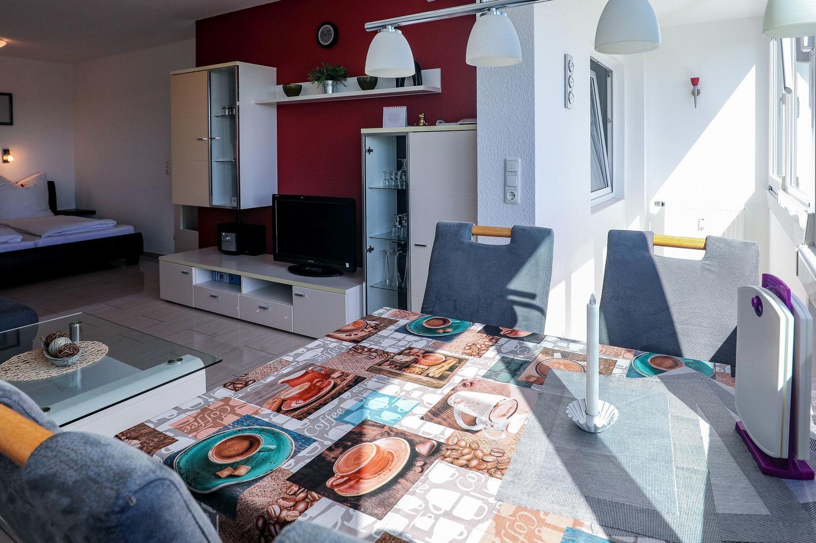 Essbereich mit Tischdecke und Stühlen. Wohnbereich mit Fernseher und Bett im Hintergrund.