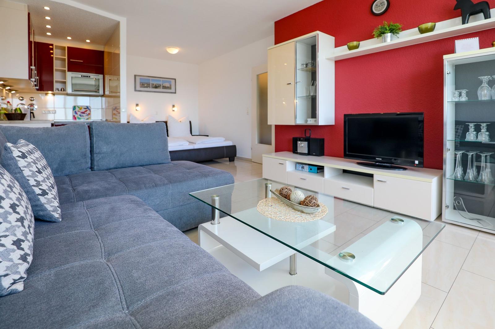 Wohnzimmer mit Sofa, Glas-Tisch und Fernseher an roter Wand.