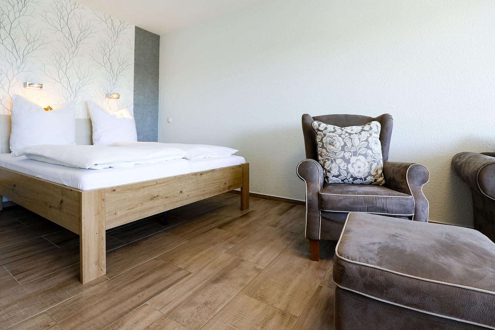 Ein Schlafzimmer mit Holzbett, Sessel und Fußhocker. Wand mit Baum-Motiv.
