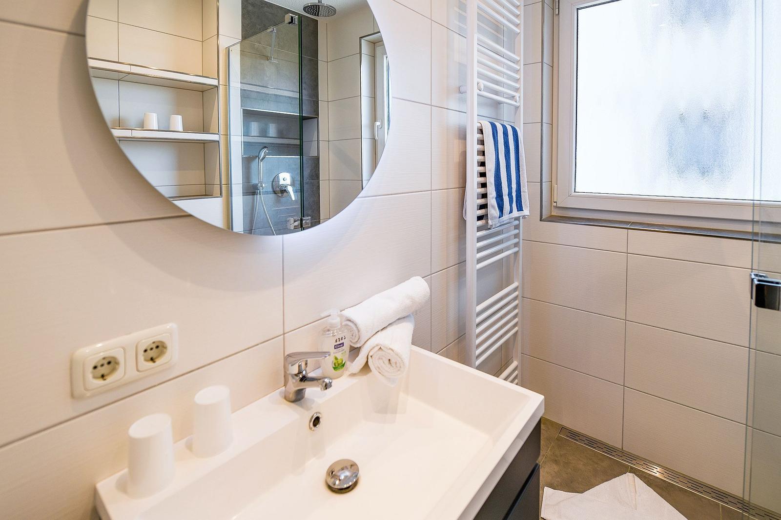 Bad mit Waschbecken, Spiegel und Dusche. Handtuchhalter und Fenster sind sichtbar.