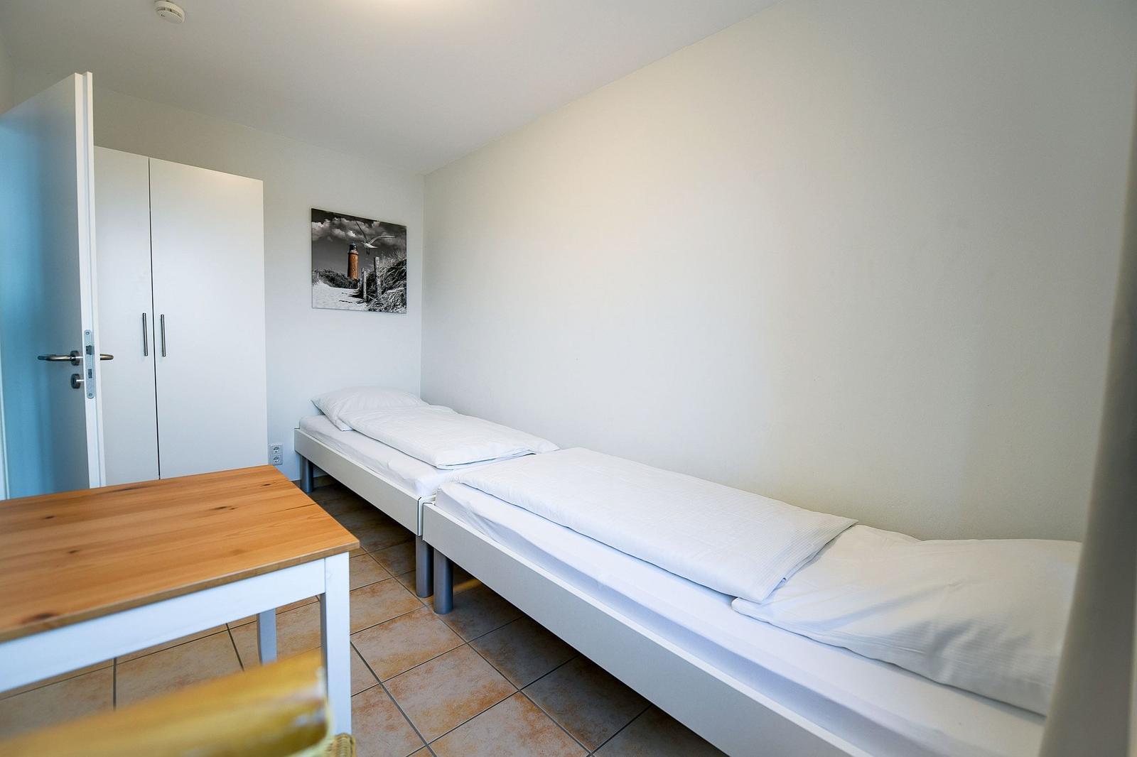 Zwei Einzelbetten, Tisch, Kleiderschrank und Wandbild in einem Zimmer.