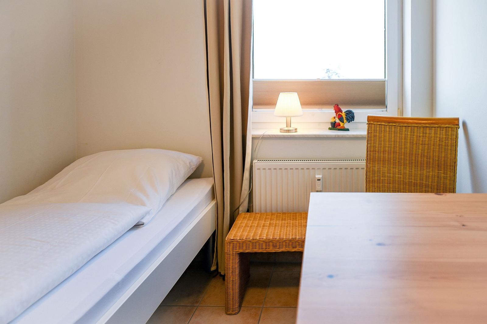 Ein Schlafzimmer mit Bett, Tisch, Stuhl und Fenster. Auf dem Fensterbrett steht eine Lampe und eine Figur.