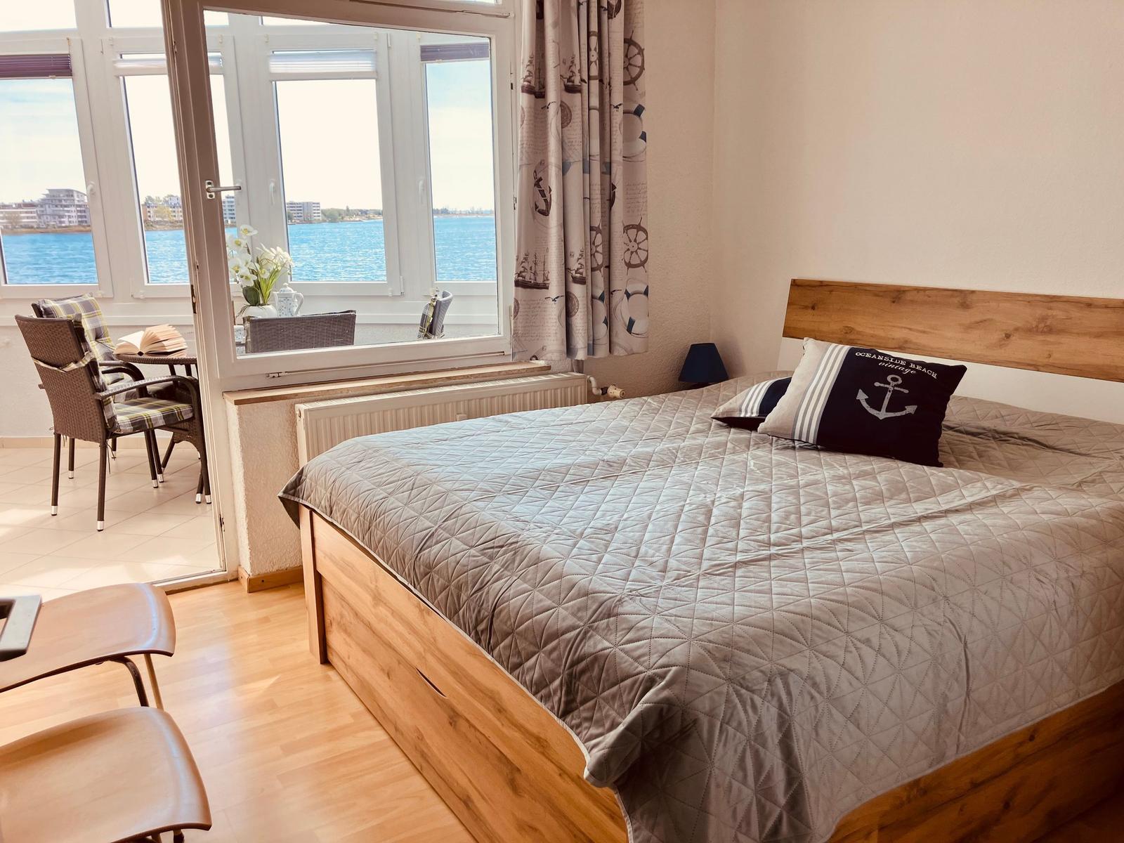 Schlafzimmer mit Blick aufs Wasser, Holzbett und Fensterbank mit Stühlen.