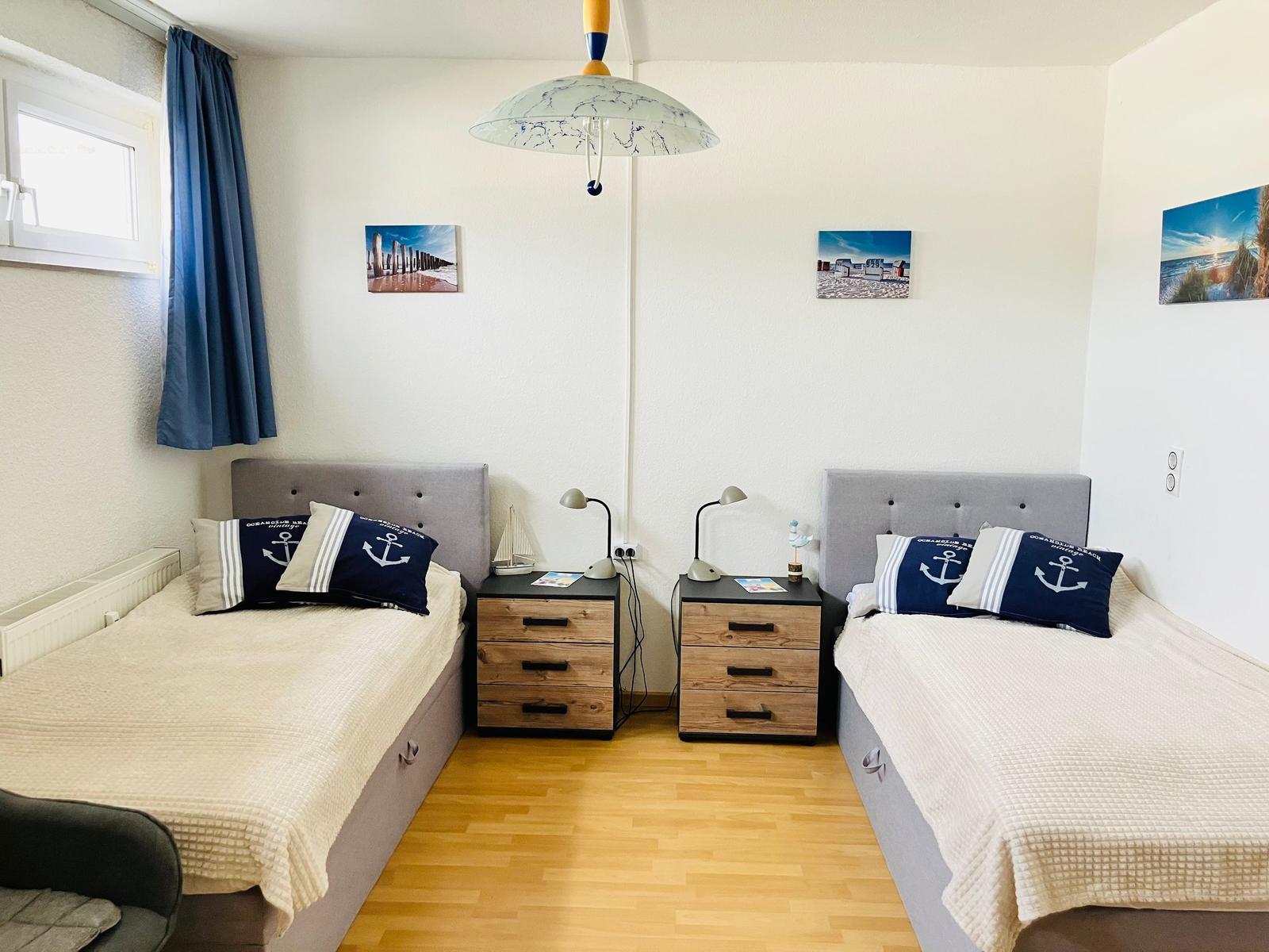 Zwei Einzelbetten mit Anker-Kissen, Nachttische und Wandbilder mit Strandmotiven.