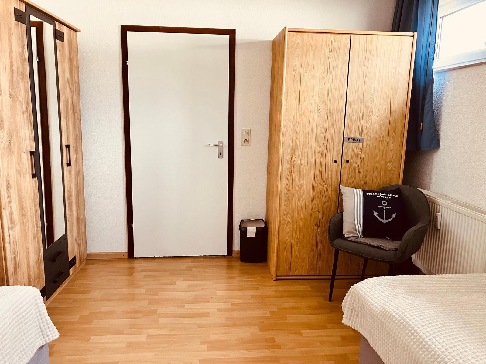 Zimmer mit zwei Schränken, Stuhl und Fenster. Holzfußboden und Tür sichtbar.