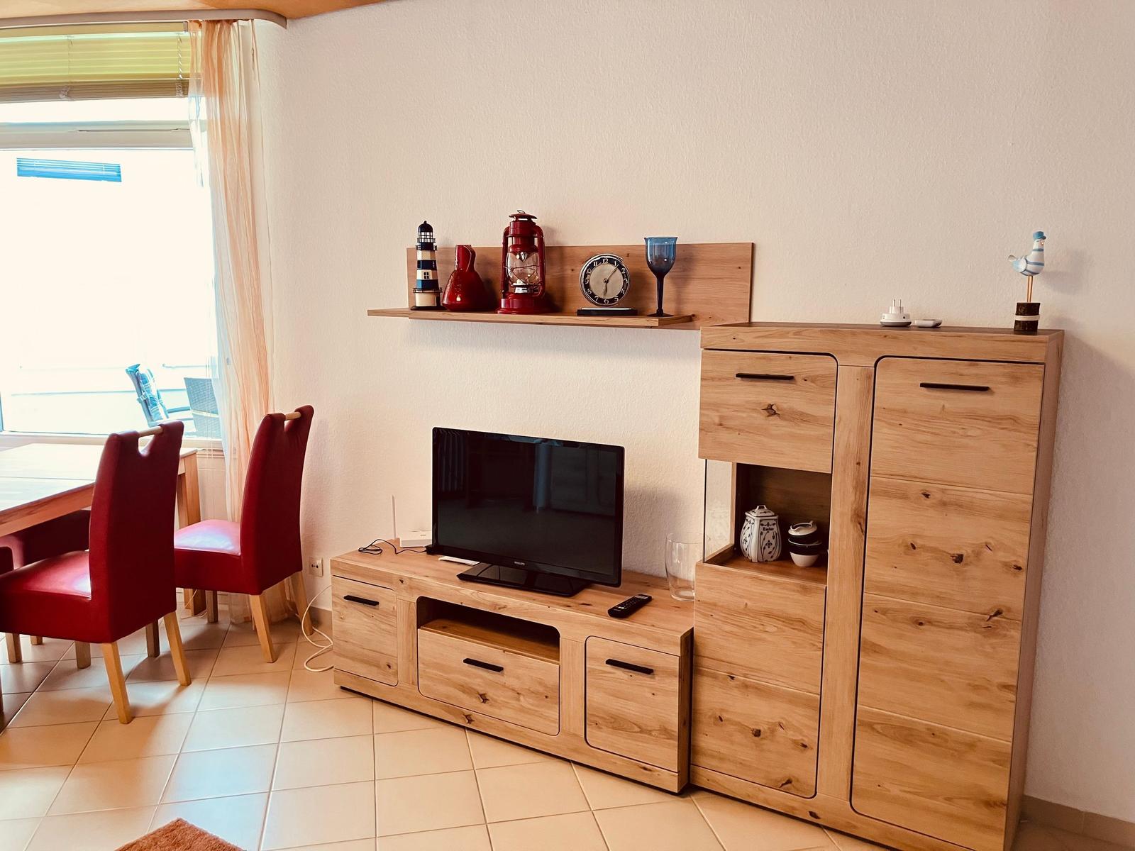 Wohnzimmer mit Holzmöbeln, Fernseher und Dekorationsgegenständen. Tisch und Stühle sind sichtbar.
