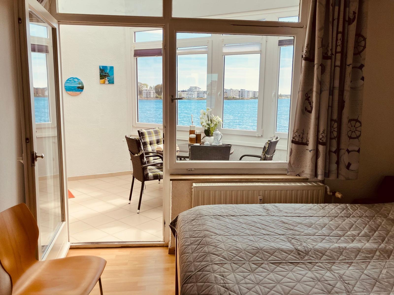 Zimmer mit Blick auf Wasser, Balkon mit Sitzgelegenheiten und Bett im Vordergrund.