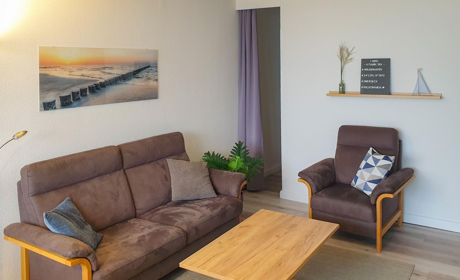 Living room with sofa, armchair, and wooden table. Wall picture shows beach. Shelf with decor.