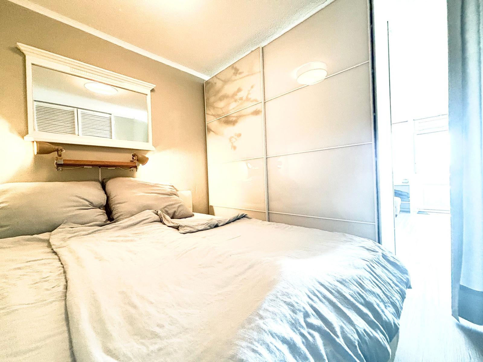 Schlafzimmer mit Bett, Spiegel und Schrankwand. Tür führt zu einem hellen Raum.