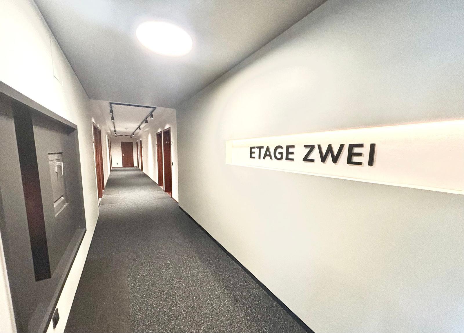 Flur mit Schild Etage Zwei und Türen. Teppichboden und Deckenbeleuchtung.