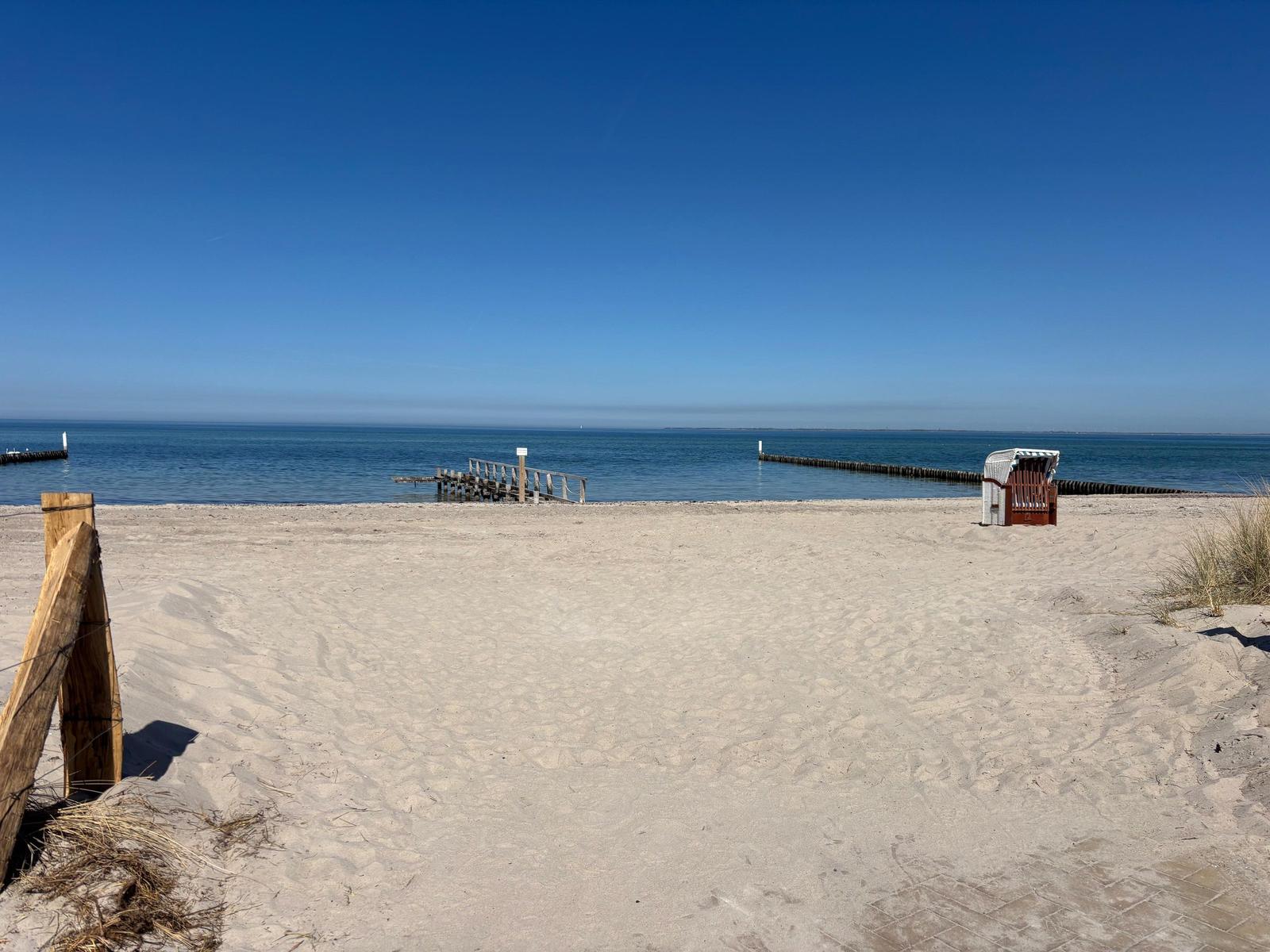 Strand mit Sand, See und Strandkorb. Holzsteg und Dünen sind sichtbar.