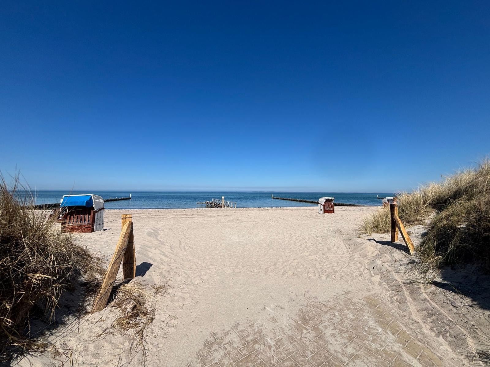 Sandweg führt zur Strandpromenade mit Strandkörben und Blick aufs Meer.