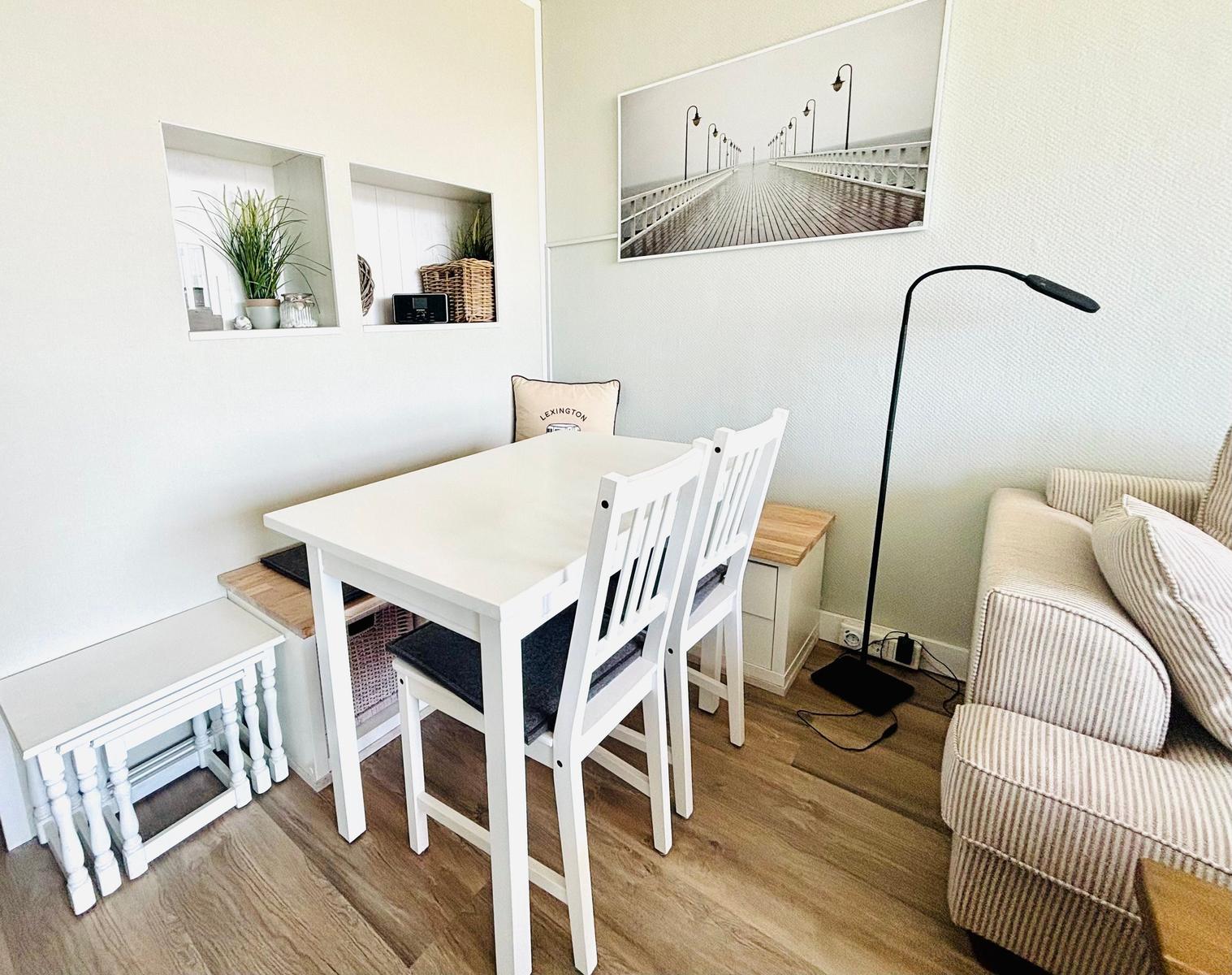 Essbereich mit weißem Tisch, Stühlen und Sofa. Wandbild und Pflanzen sind sichtbar.