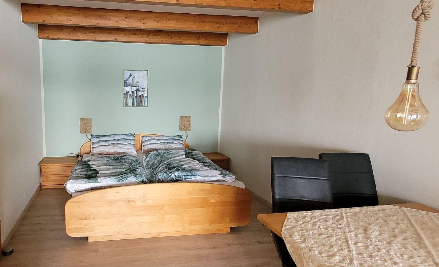 Schlafzimmer mit Holzbett, Nachttischen und Decke. An der Wand hängt ein Bild. Hängelampe und Tisch mit Stühlen.