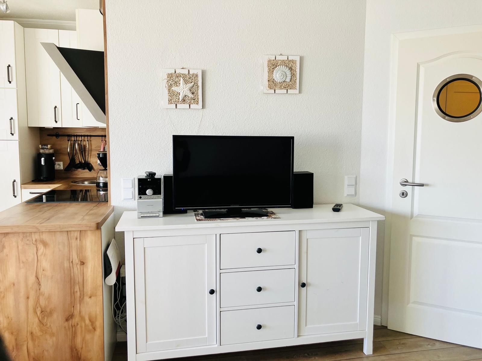 TV auf weißen Schrank, Küchenbereich links, Tür rechts mit runden Fenster.