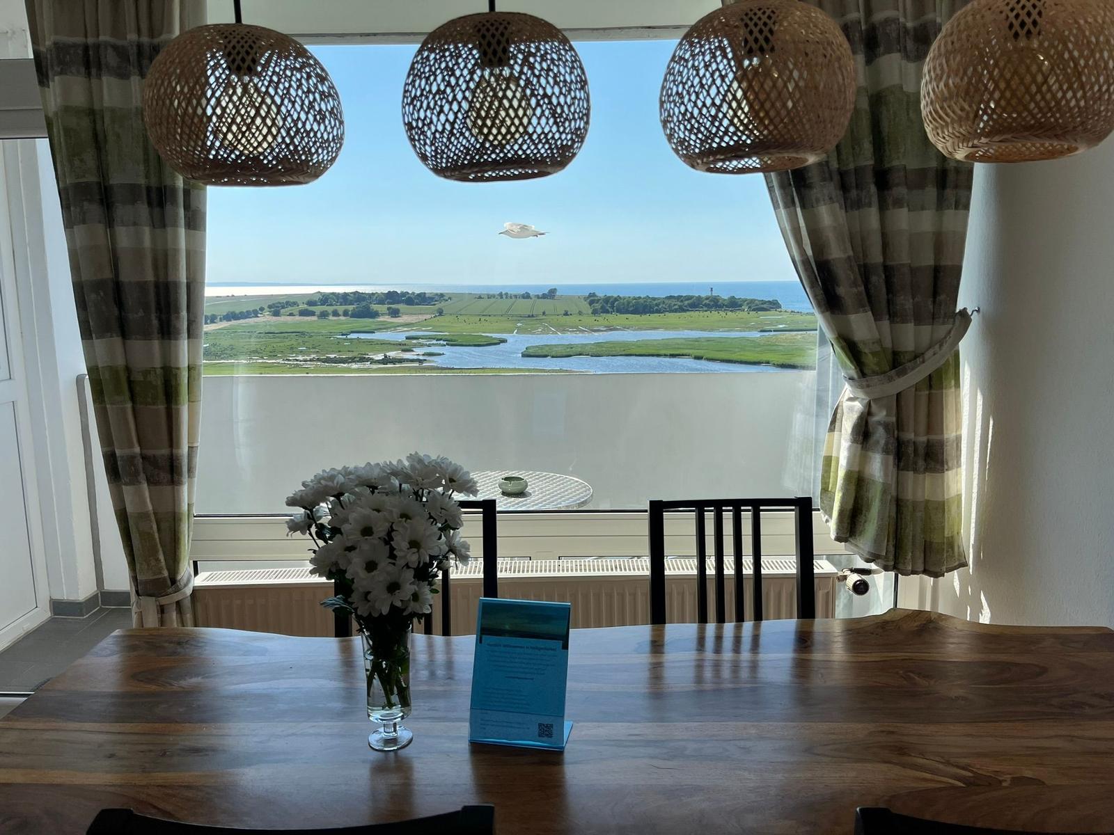 Essbereich mit Blick auf Fluss und Landschaft. Holztisch, Blumen und Hängelampen.