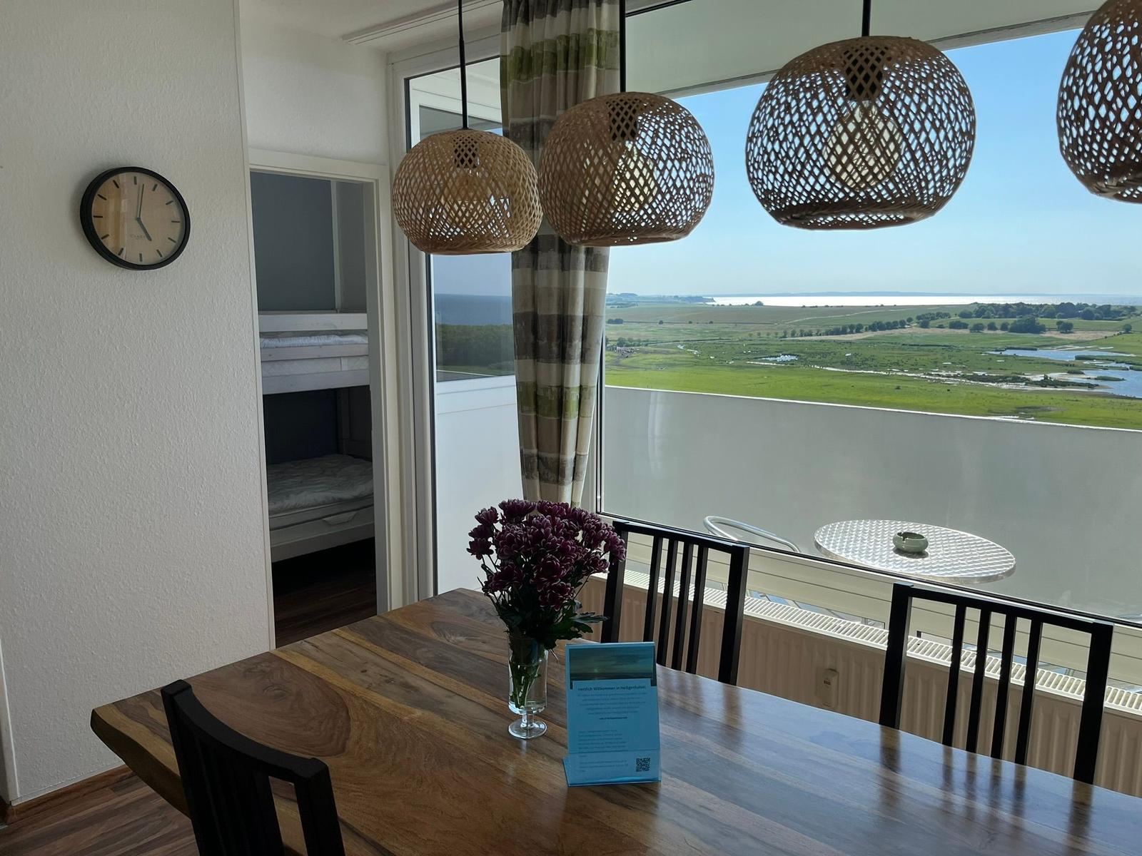 Essbereich mit Blick auf Landschaft und Meer. Holztisch, Stühle und hängende Lampen.