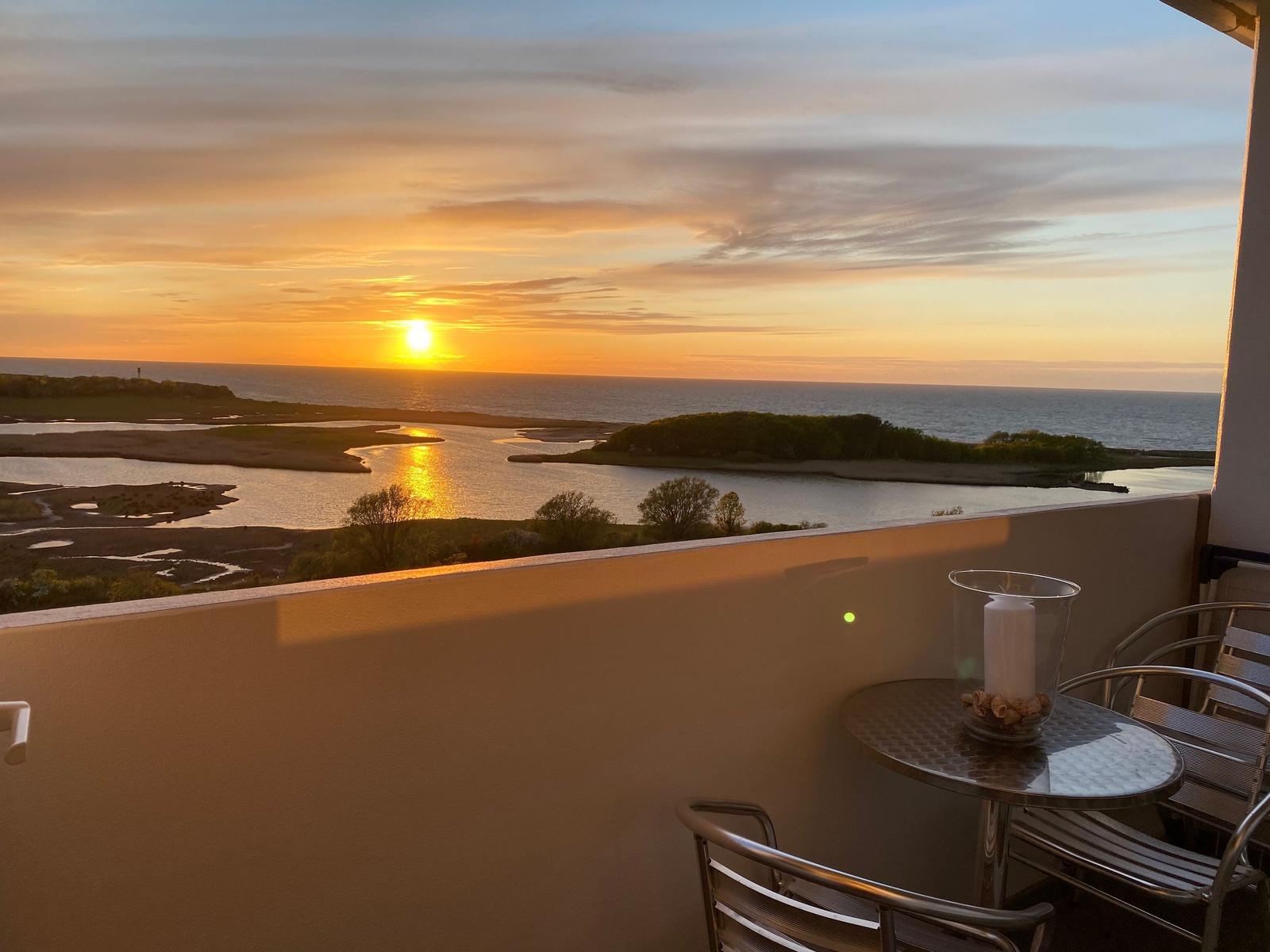 Terrasse mit Tisch und Stühlen bietet Blick auf Sonnenuntergang über Wasser und Inseln.