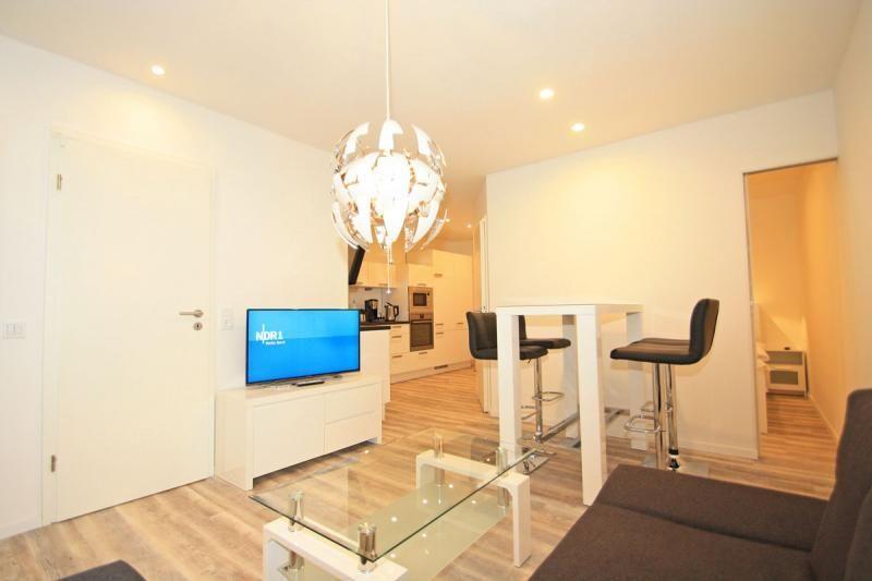 Living area with TV, glass table, and bar stools. Kitchen and bedroom are visible.