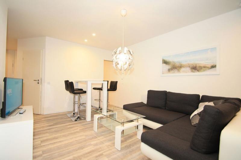 Living room with sofa, glass table, and bar stools. Wall art and ceiling light visible.