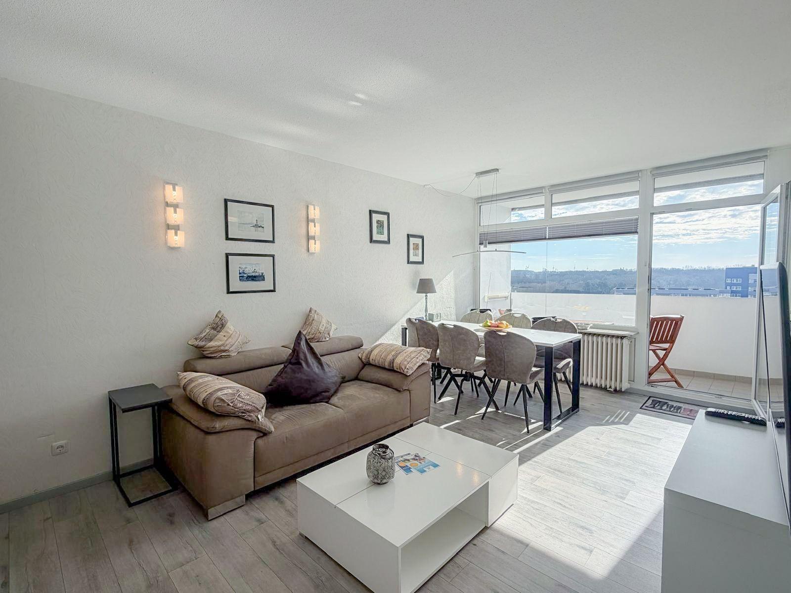 Wohn- und Essbereich mit Sofa, Tisch und Sitzgelegenheiten. Blick auf Balkon und Stadtsilhouette.