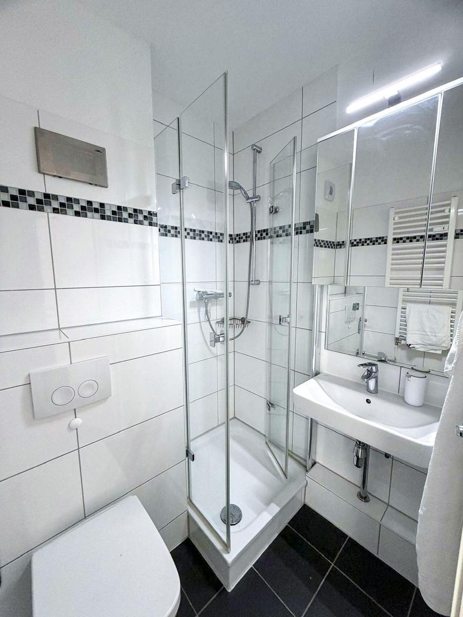 Badezimmer mit Dusche, Waschbecken und Toilette. Weiße Fliesen und schwarze Bodenfliesen.