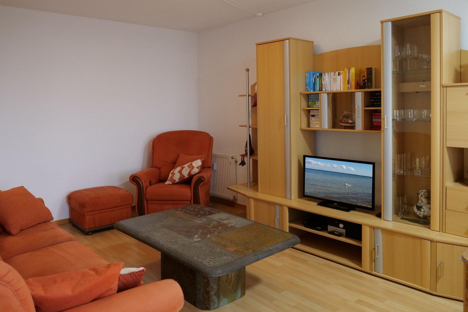 Living room with orange sofa, armchair, and wooden furniture. TV shows sea view.