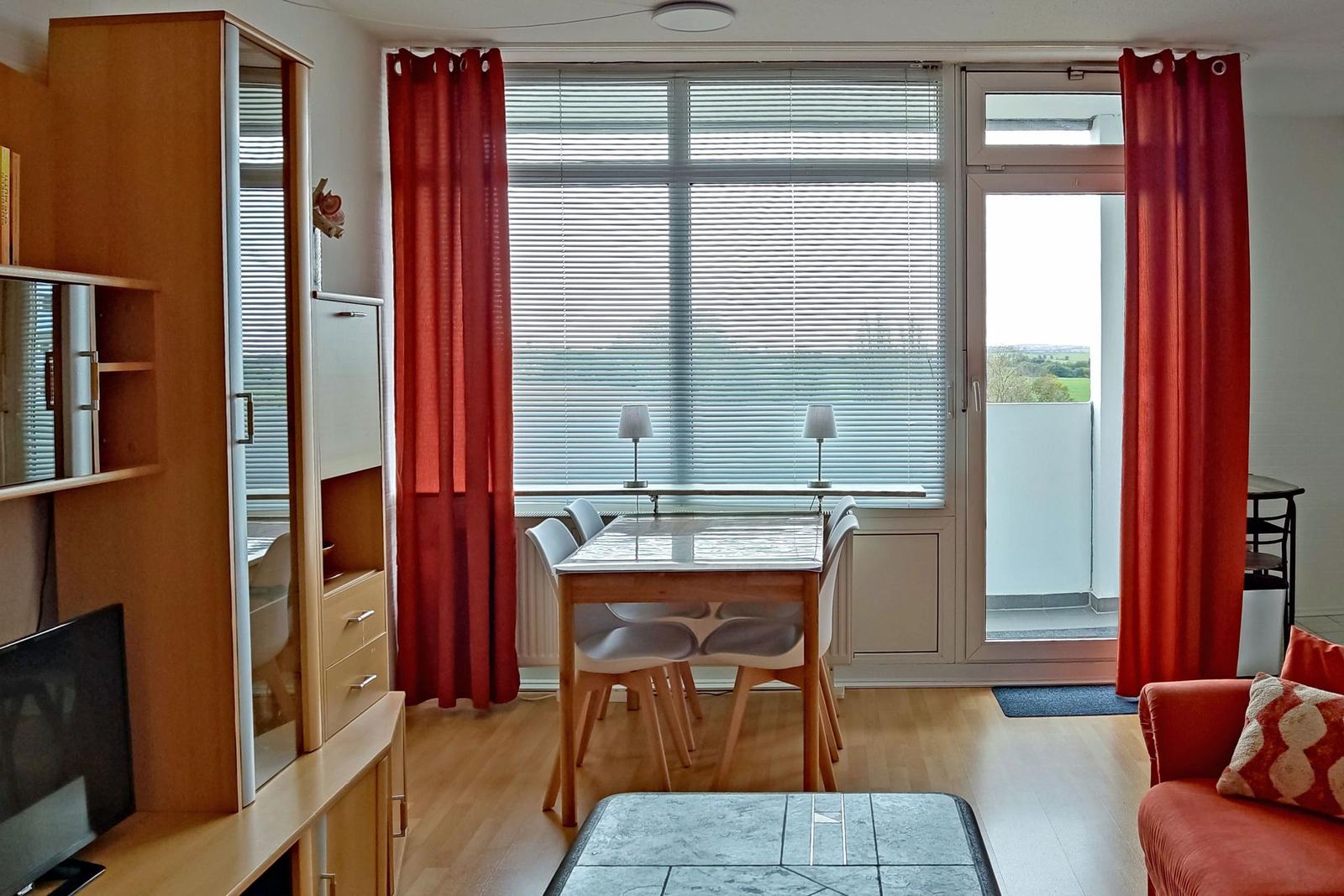 Dining area with table, chairs, and view to balcony. Red curtains and wooden floor.