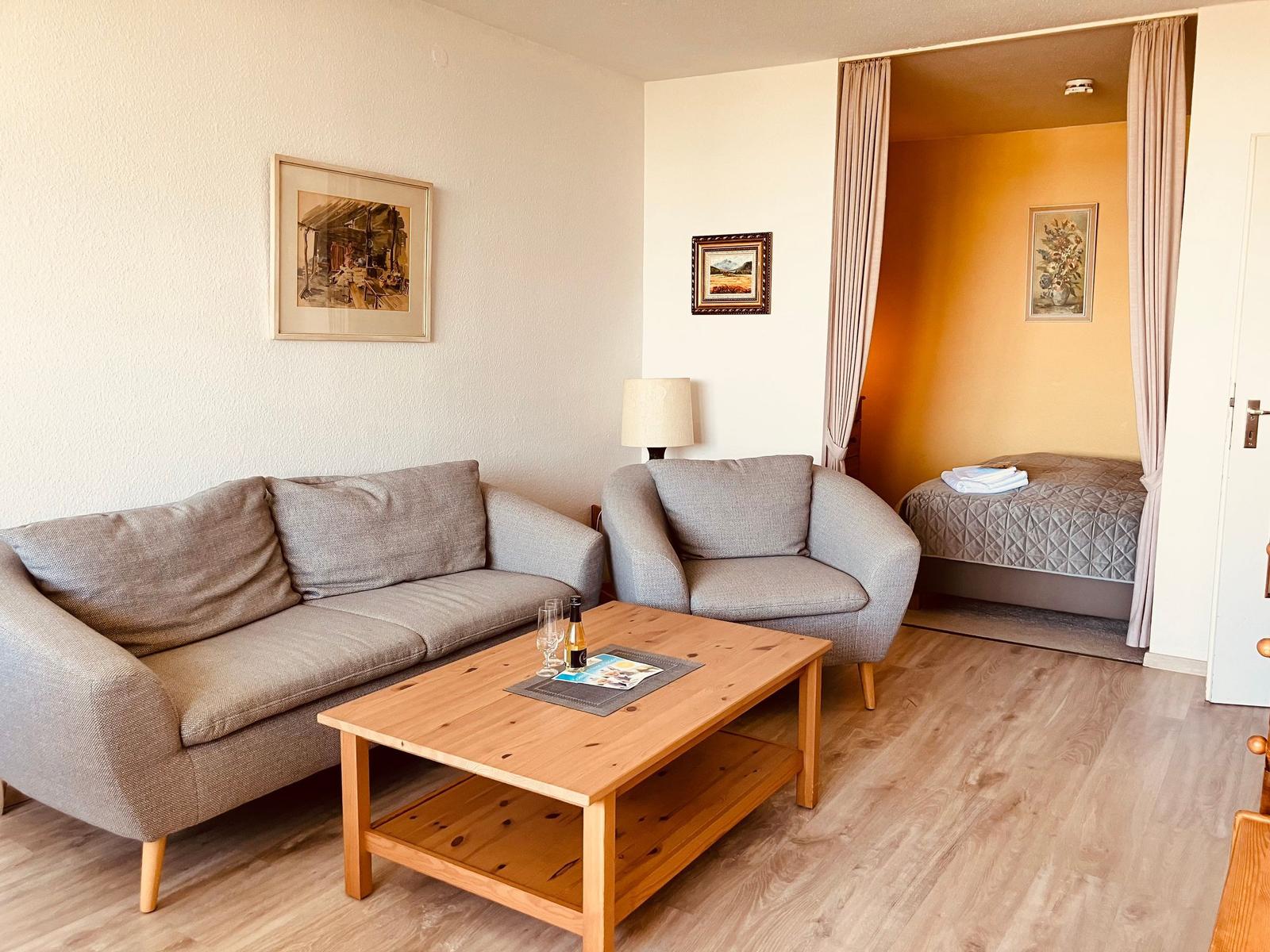 Living area with sofa, armchair, and wooden table. Bedroom with bed visible in background.