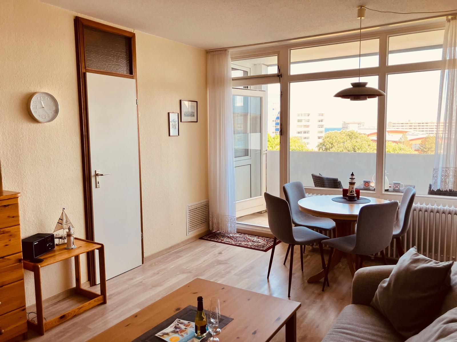 Living area with dining space, balcony, and wooden floor. Windows overlook the city.
