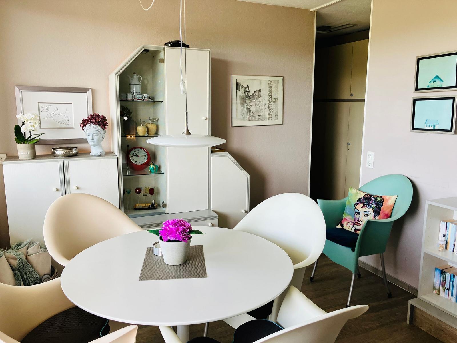Runde weiße Tisch mit Stühlen, Vitrine, Bilder und grüner Sessel mit Kissen.