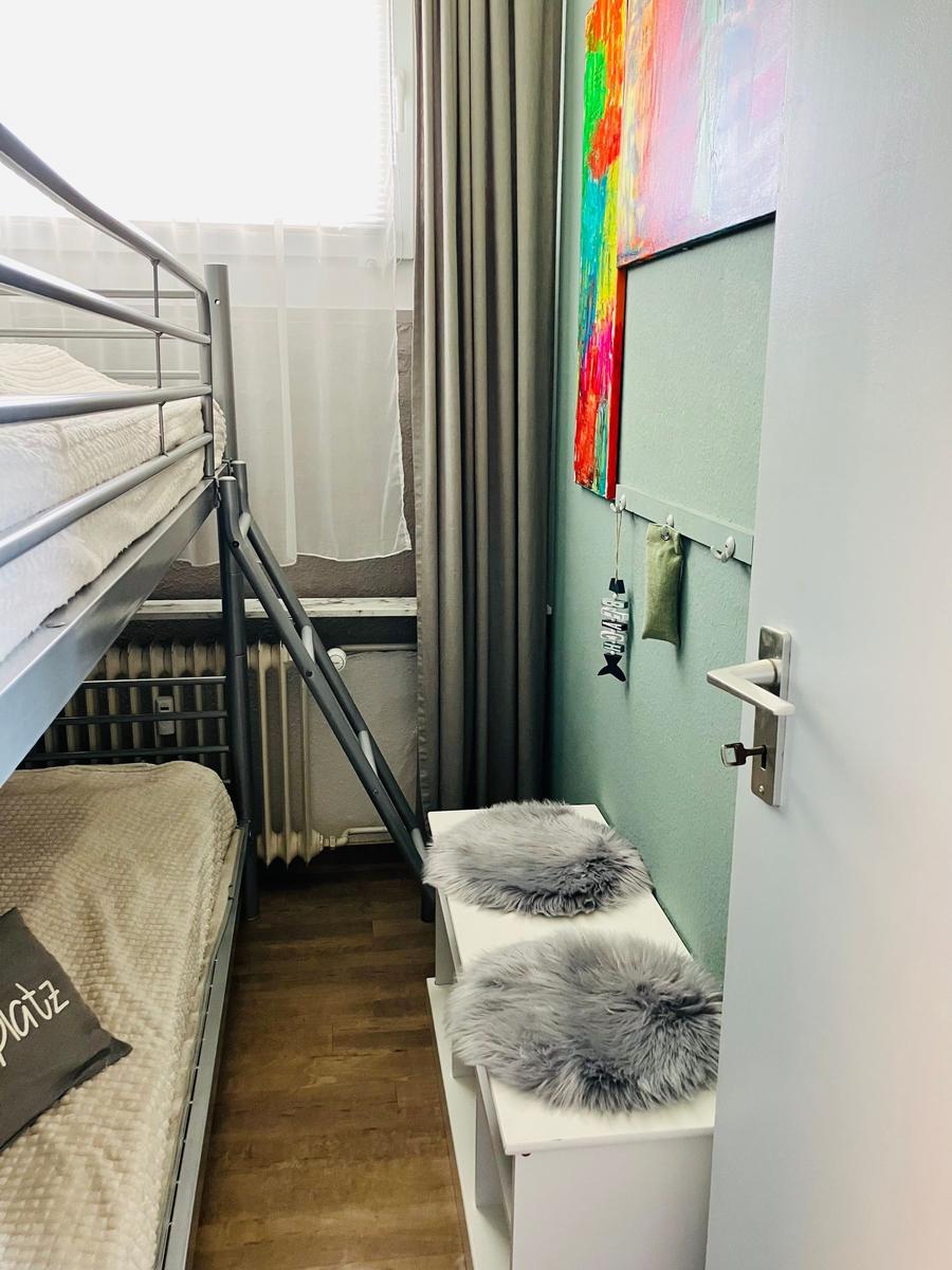 Zimmer mit Doppelstockbett, Fenster, Wandbild und grauen Fellkissen auf einem weißen Regal.