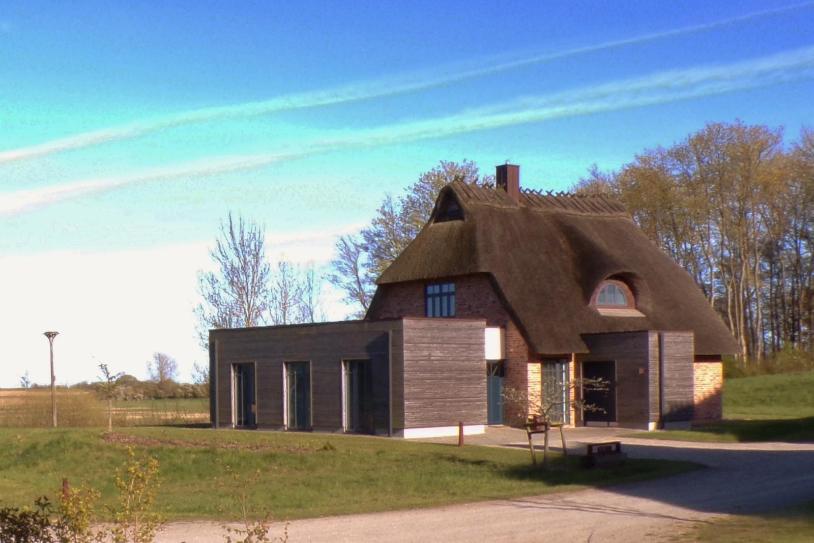 Haus mit Strohdach und Holzfassade, umgeben von Gras und Bäumen unter blauem Himmel.