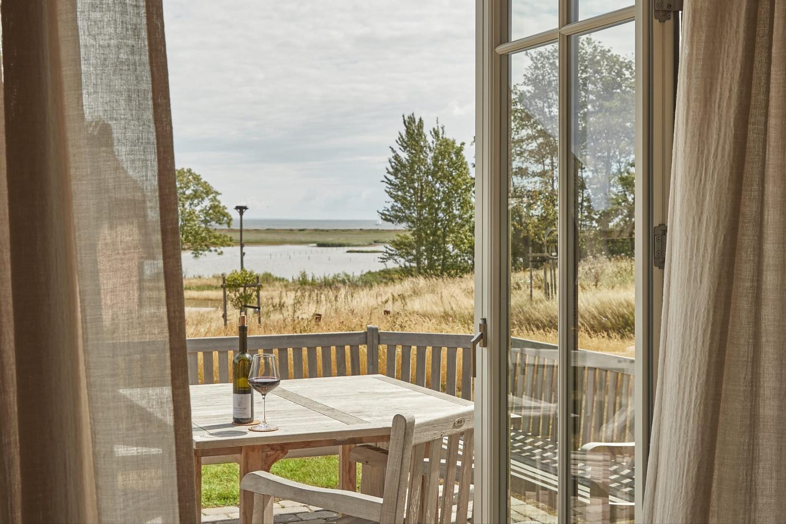 Durch die Glastür blickt man auf eine Terrasse mit Tisch, Stühlen und Blick auf Wasser und Bäume.