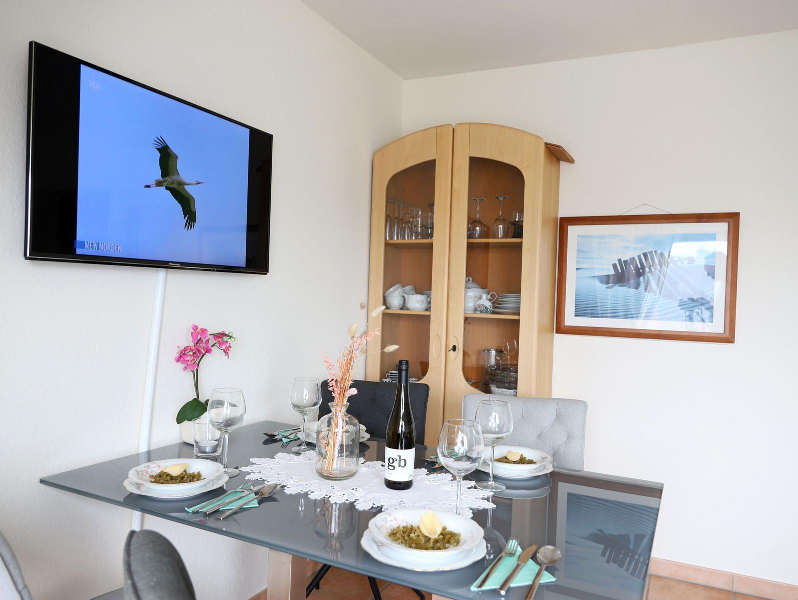 Esstisch mit Platz für zwei, Fernseher und Glasvitrine im Hintergrund.