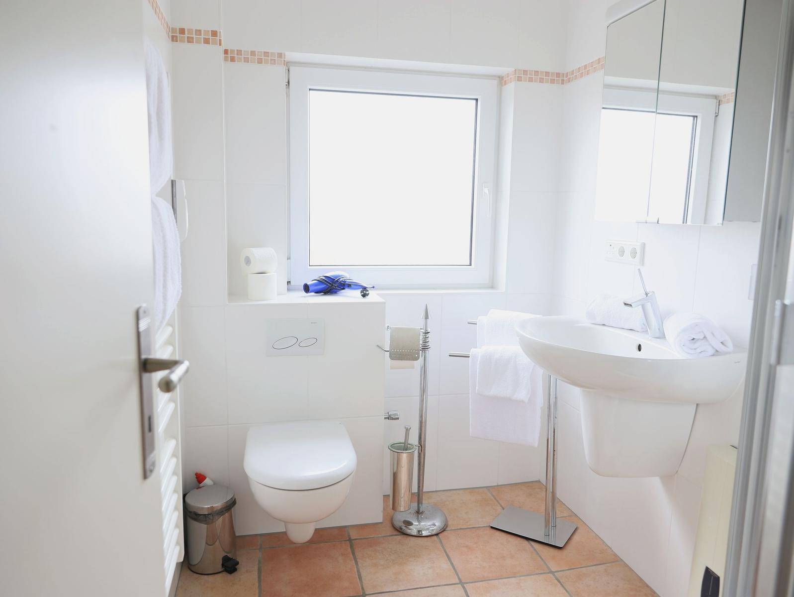 Badezimmer mit Toilette, Waschbecken und Fenster. Weiße Fliesen und braune Bodenfliesen.