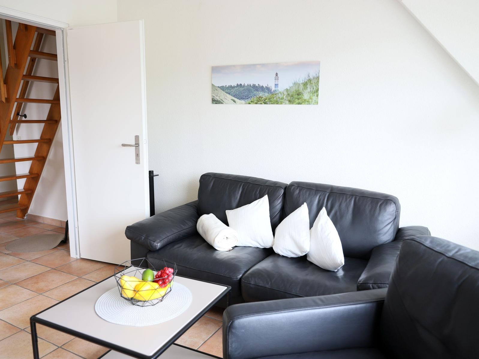 Schwarzes Sofa mit weißen Kissen, Kaffeetisch mit Obst, Wandbild und Holztreppe.
