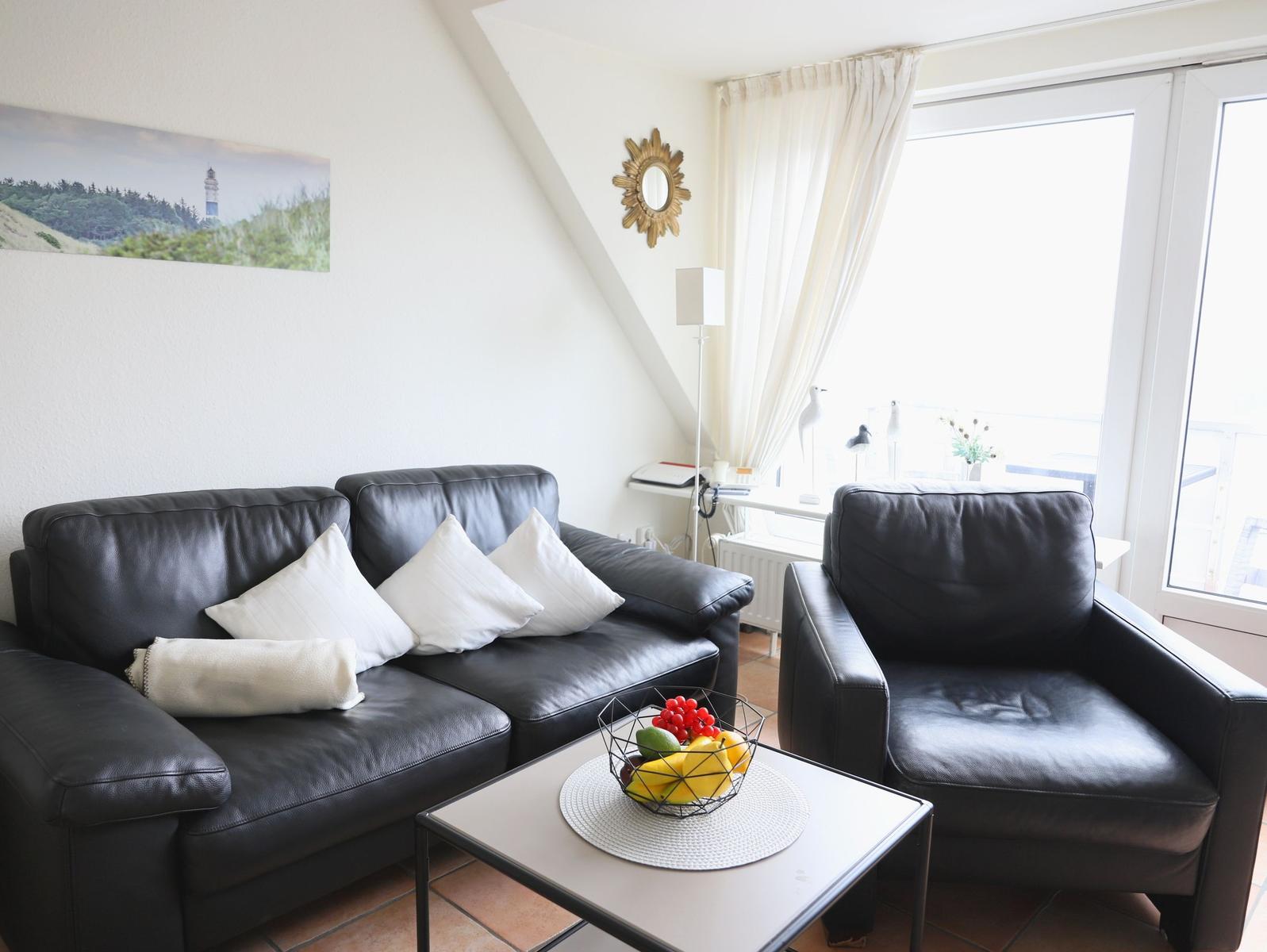 Schwarzes Sofa und Sessel mit weißen Kissen. Tisch mit Obst. Fenster mit Blick.