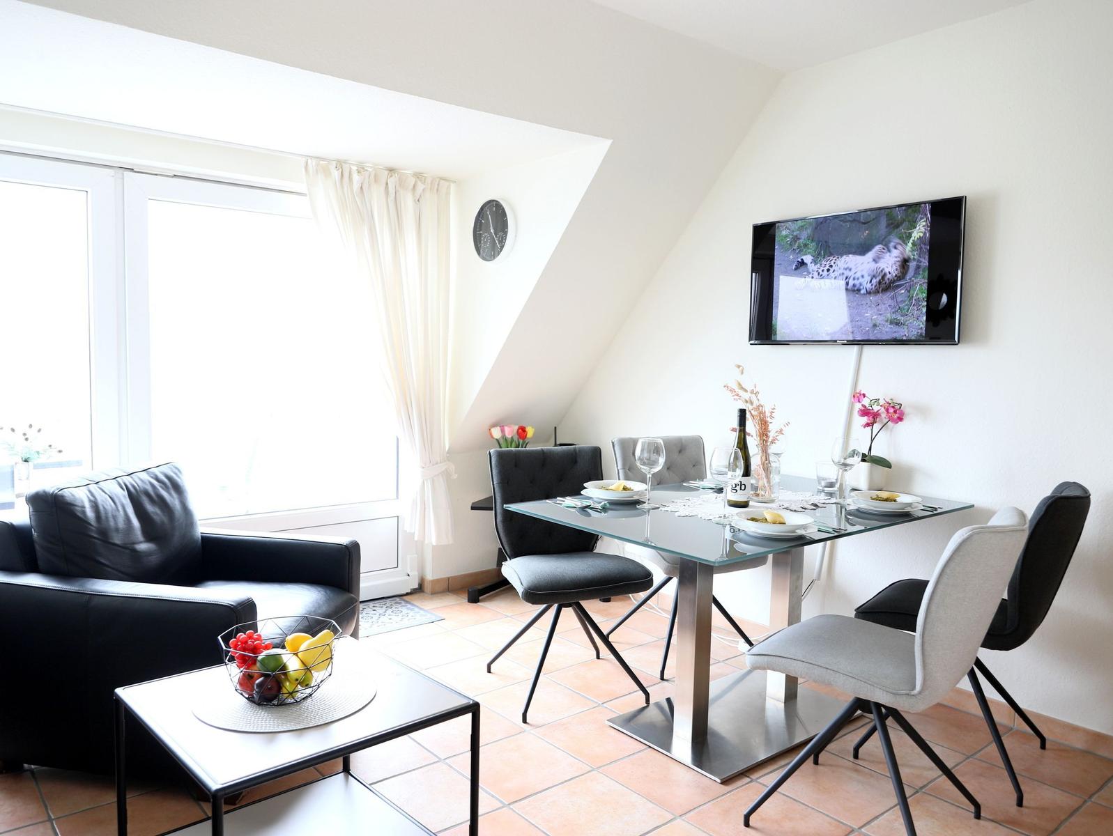 Essbereich mit Glas-Tisch und Stühlen, Fernseher an Wand, Sofa und Fenster mit Vorhängen.