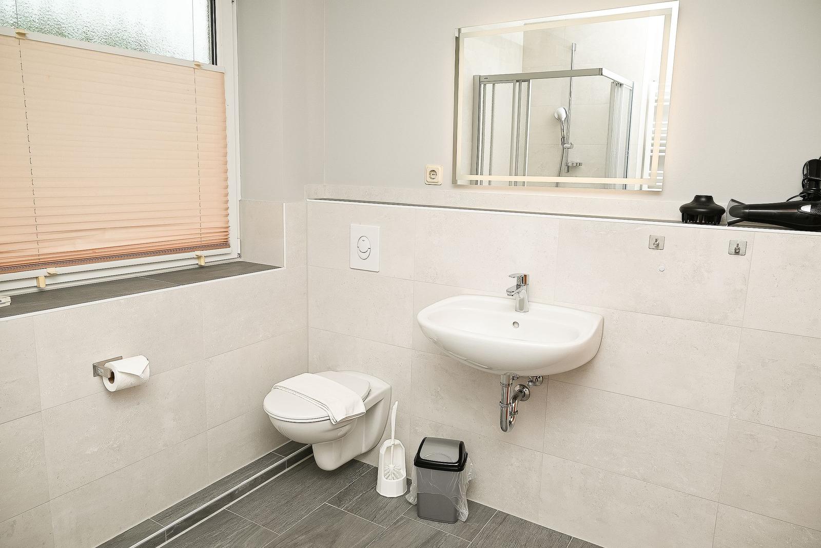 Badezimmer mit Toilette, Waschbecken und Spiegel. Fenster mit Jalousie und Duschbereich sichtbar.